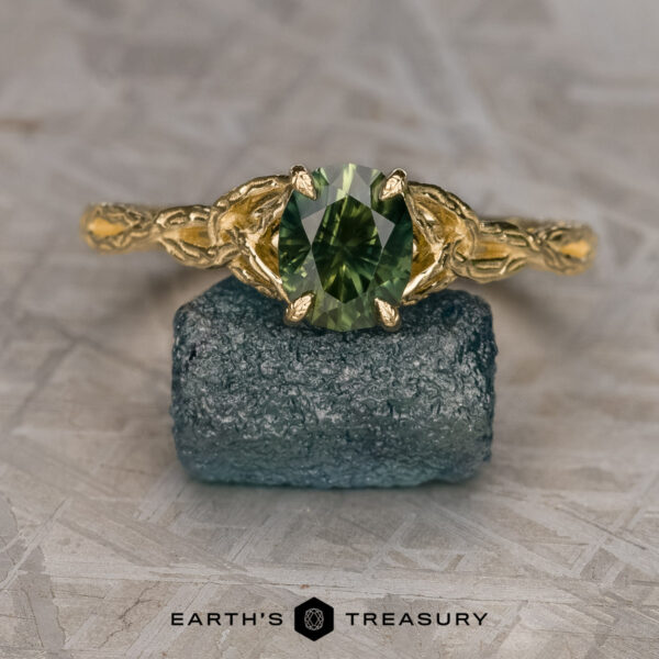 The "Guinevere" ring in 14k yellow gold with 1.41-carat Australian sapphire The "Guinevere" ring in 14k yellow gold with a 1.41-carat Australian sapphire from the Guinevere Solitaire collection is shown on a rough blue stone, with the "Earth's Treasury" logo and text at the bottom.