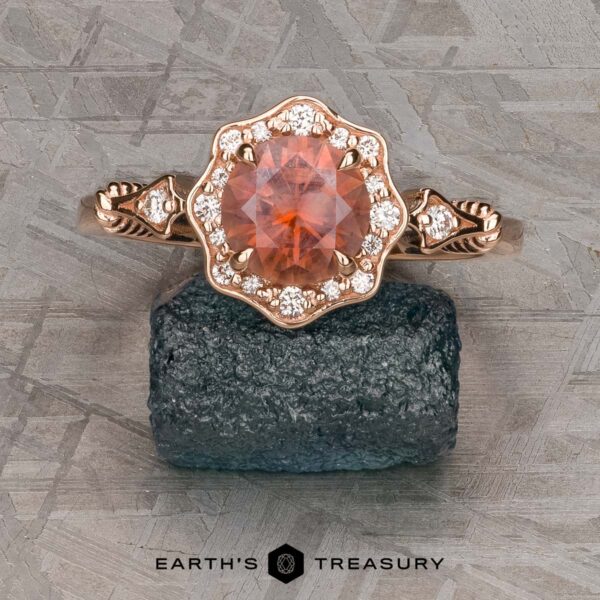 The "Cordelia" ring in 14K rose gold with 1.38-carat Montana sapphire The "Cordelia" ring in 14K rose gold showcases a 1.38-carat Montana sapphire surrounded by small diamonds, set upright on a dark mineral rock with “Earth’s Treasury” branding below.
