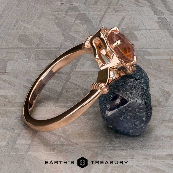 The "Cordelia" ring in 14K rose gold with 1.38-carat Montana sapphire The "Cordelia" ring in 14K rose gold showcases a 1.38-carat Montana sapphire surrounded by small diamonds, set upright on a dark mineral rock with “Earth’s Treasury” branding below.