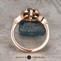 The "Cordelia" ring in 14K rose gold showcases a 1.38-carat Montana sapphire surrounded by small diamonds, set upright on a dark mineral rock with “Earth’s Treasury” branding below.
