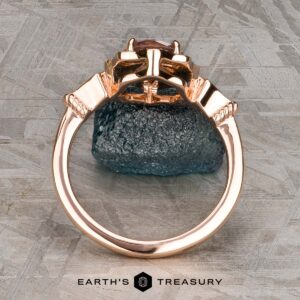 The "Cordelia" ring in 14K rose gold showcases a 1.38-carat Montana sapphire surrounded by small diamonds, set upright on a dark mineral rock with “Earth’s Treasury” branding below.