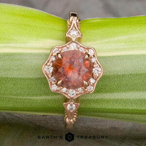 The "Cordelia" ring in 14K rose gold showcases a 1.38-carat Montana sapphire surrounded by small diamonds, set upright on a dark mineral rock with “Earth’s Treasury” branding below.