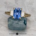 The "Damara" ring in 18k yellow gold showcases a 2.44-carat Madagascar sapphire center stone, accented by two clear stones, elegantly displayed atop a rough blue mineral on a textured gray surface.