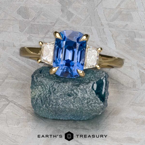 The "Damara" ring in 18k yellow gold with 2.44-carat Madagascar sapphire The "Damara" ring in 18k yellow gold showcases a 2.44-carat Madagascar sapphire center stone, accented by two clear stones, elegantly displayed atop a rough blue mineral on a textured gray surface.
