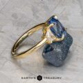 The "Damara" ring in 18k yellow gold showcases a 2.44-carat Madagascar sapphire center stone, accented by two clear stones, elegantly displayed atop a rough blue mineral on a textured gray surface.