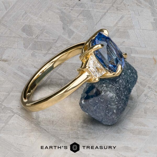 The "Damara" ring in 18k yellow gold with 2.44-carat Madagascar sapphire The "Damara" ring in 18k yellow gold showcases a 2.44-carat Madagascar sapphire center stone, accented by two clear stones, elegantly displayed atop a rough blue mineral on a textured gray surface.