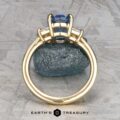 The "Damara" ring in 18k yellow gold showcases a 2.44-carat Madagascar sapphire center stone, accented by two clear stones, elegantly displayed atop a rough blue mineral on a textured gray surface.