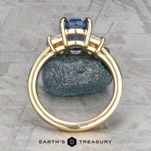 The "Damara" ring in 18k yellow gold showcases a 2.44-carat Madagascar sapphire center stone, accented by two clear stones, elegantly displayed atop a rough blue mineral on a textured gray surface.