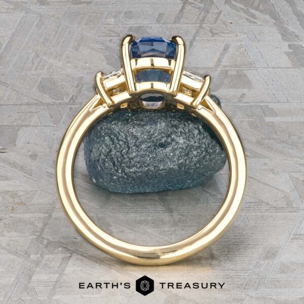 The "Damara" ring in 18k yellow gold with 2.44-carat Madagascar sapphire The "Damara" ring in 18k yellow gold showcases a 2.44-carat Madagascar sapphire center stone, accented by two clear stones, elegantly displayed atop a rough blue mineral on a textured gray surface.