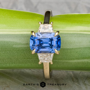 The "Damara" ring in 18k yellow gold showcases a 2.44-carat Madagascar sapphire center stone, accented by two clear stones, elegantly displayed atop a rough blue mineral on a textured gray surface.