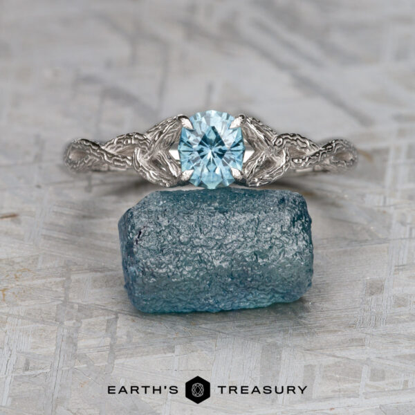 The "Guinevere" ring in 14K white gold with 0.84-carat Montana sapphire The "Guinevere" ring in 14k white gold with a 0.84-carat Montana sapphire rests atop a rough blue crystal on a textured gray surface, with the text "EARTH'S TREASURY" displayed below.