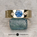 The "Hermes" ring in hammered 14k yellow gold features a 0.99-carat blue gemstone at its center, flanked by two diamonds, and is elegantly displayed on a rough blue-green stone.