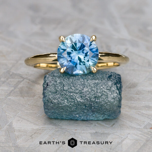 The "Ripple" ring in 18K yellow gold with 1.90-carat Montana sapphire The "Ripple" ring in 18k yellow gold with a 1.90-carat Montana sapphire rests atop a rough blue crystal, with the "Earth's Treasury" logo shown below.