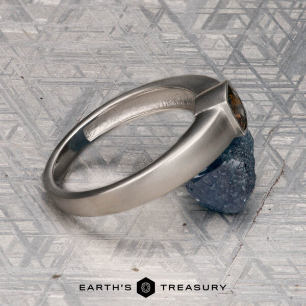 The "Declan " ring in platinum with 1.02-carat Montana sapphire The "Declan" ring in platinum with a 1.02-carat Montana sapphire rests on a rough blue crystal atop a patterned metallic surface, with the "EARTH'S TREASURY" logo shown at the bottom.