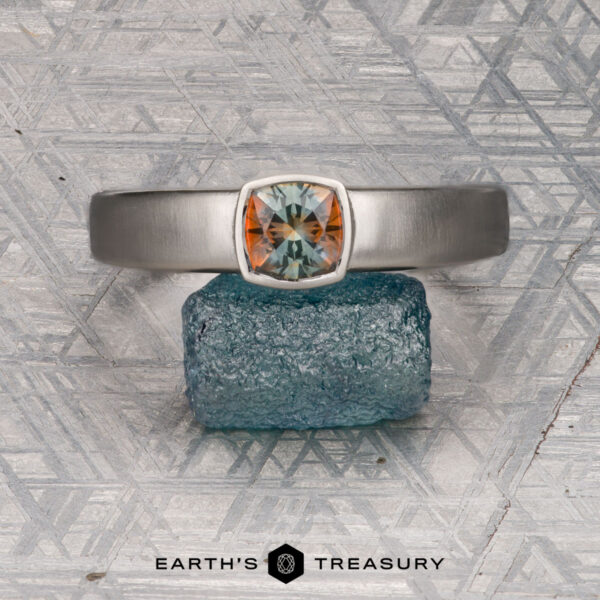 The "Declan " ring in platinum with 1.02-carat Montana sapphire The "Declan" ring in platinum, featuring a 1.02-carat Montana sapphire, is displayed atop a rough blue stone against a geometric-patterned background. "EARTH'S TREASURY" appears at the bottom.