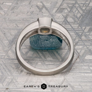 The "Declan" ring in platinum, featuring a 1.02-carat Montana sapphire, stands upright on a textured surface with blue glass beneath it. The Earth's Treasury logo is displayed at the bottom.