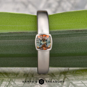 A platinum "Declan" ring with a 1.02-carat Montana sapphire, featuring vibrant orange and green hues, displayed on a green leaf with "EARTH'S TREASURY" below.