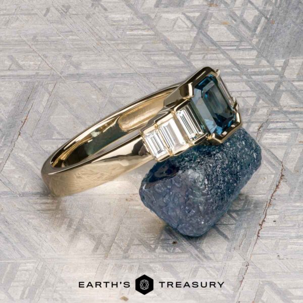 The "Apollo" ring in 14K yellow gold with 1.35-carat Montana sapphire The "Apollo" ring in 14K yellow gold with a 1.35-carat Montana sapphire is displayed on a rough crystal, with the "Earth's Treasury" logo below.