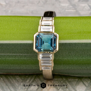 The "Apollo" ring features a 1.35-carat Montana sapphire set in 14K yellow gold, displayed elegantly on a green and silver background.
