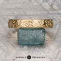An 18K yellow gold Hawthorne band is displayed on a blue textured stone next to a gold floral bracelet, with "EARTH'S TREASURY" written below on a geometric-patterned surface.