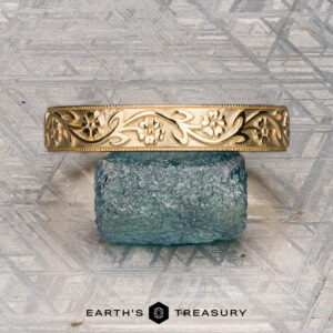 An 18K yellow gold Hawthorne band is displayed on a blue textured stone next to a gold floral bracelet, with "EARTH'S TREASURY" written below on a geometric-patterned surface.