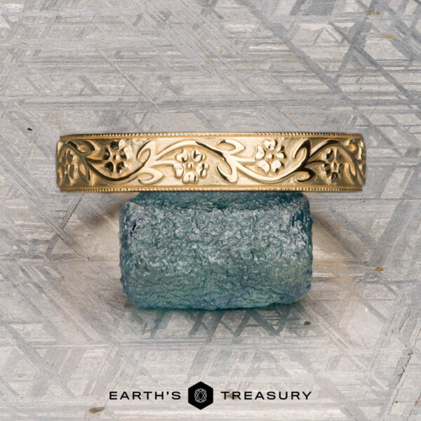 An 18K yellow gold Hawthorne band is displayed on a blue textured stone next to a gold floral bracelet, with "EARTH'S TREASURY" written below on a geometric-patterned surface.