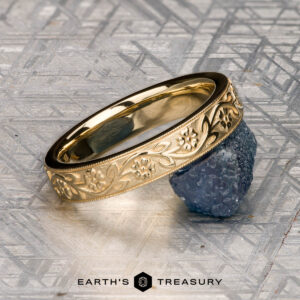 An 18K yellow gold Hawthorne band is displayed on a blue textured stone next to a gold floral bracelet, with "EARTH'S TREASURY" written below on a geometric-patterned surface.