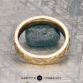 An 18K yellow gold Hawthorne band is displayed on a blue textured stone next to a gold floral bracelet, with "EARTH'S TREASURY" written below on a geometric-patterned surface.