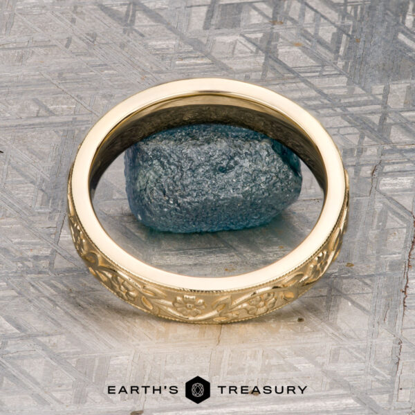 The "Hawthorne" band in 18K yellow gold An 18K yellow gold Hawthorne band is displayed on a blue textured stone next to a gold floral bracelet, with "EARTH'S TREASURY" written below on a geometric-patterned surface.