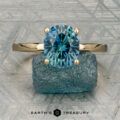 The "Aubrey" ring in 14K yellow gold, featuring a 3.37-carat Montana sapphire and hidden halo, is showcased on a rough blue stone atop a textured gray surface with the "Earth’s Treasury" logo below.