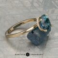 The "Aubrey" ring in 14K yellow gold, featuring a 3.37-carat Montana sapphire and hidden halo, is showcased on a rough blue stone atop a textured gray surface with the "Earth’s Treasury" logo below.