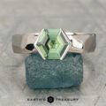 The "Ezra" ring in 14K white gold featuring a 1.64-carat Madagascar sapphire is displayed upright on a rough green-blue stone, with "Earth's Treasury" text below.