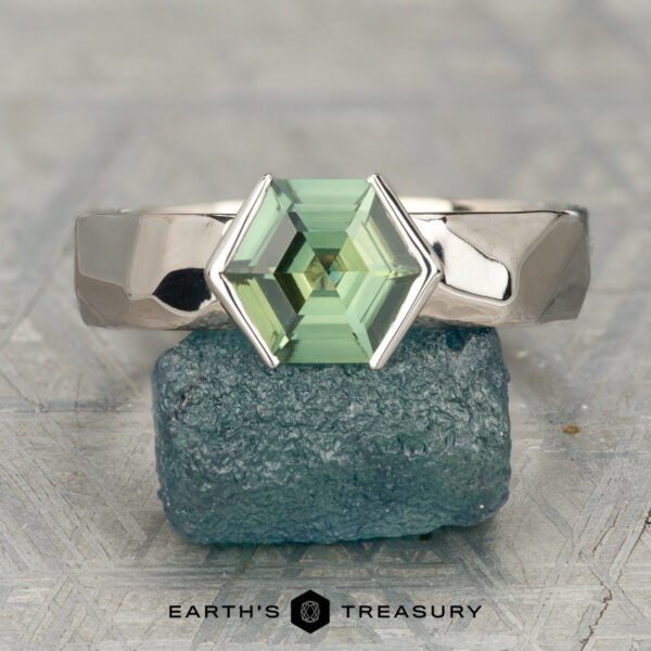 The "Ezra" ring in 14K white gold with 1.64-carat Madagascar sapphire The "Ezra" ring in 14K white gold featuring a 1.64-carat Madagascar sapphire is displayed upright on a rough green-blue stone, with "Earth's Treasury" text below.