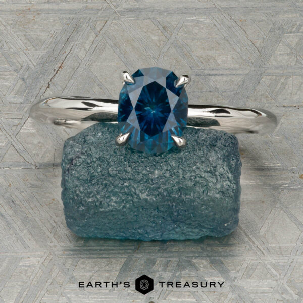 The "Ripple" ring in platinum with 1.23-carat Montana sapphire The "Ripple" ring in platinum, featuring a 1.23-carat Montana sapphire, is shown on a rough blue stone with a patterned gray background. "EARTH'S TREASURY" is displayed at the bottom.