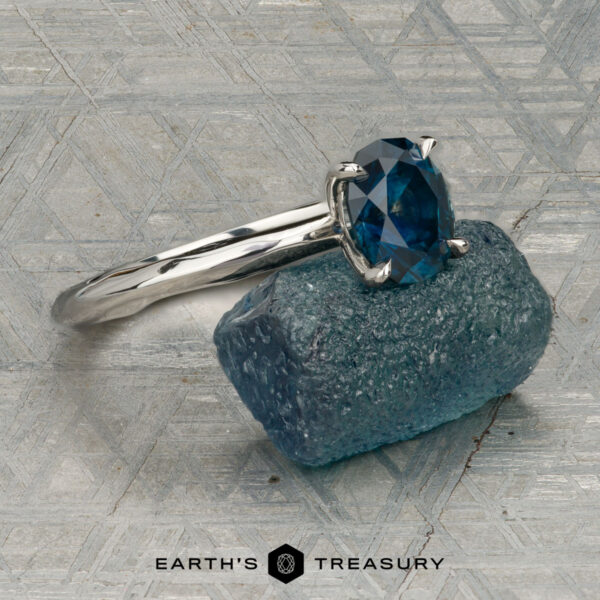The "Ripple" ring in platinum with 1.23-carat Montana sapphire The "Ripple" ring in platinum, featuring a 1.23-carat Montana sapphire, is shown on a rough blue stone with a patterned gray background. "EARTH'S TREASURY" is displayed at the bottom.