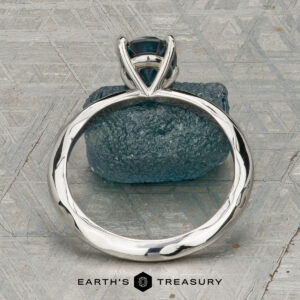 The "Ripple" ring in platinum, featuring a 1.23-carat Montana sapphire, is shown on a rough blue stone with a patterned gray background. "EARTH'S TREASURY" is displayed at the bottom.