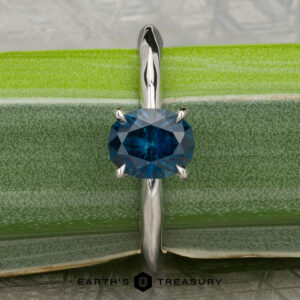 A platinum "Ripple" ring featuring a 1.23-carat Montana sapphire stands upright on a green leaf, with the Earth’s Treasury logo at the bottom.