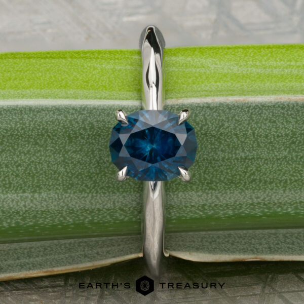 The "Ripple" ring in platinum with 1.23-carat Montana sapphire A platinum "Ripple" ring featuring a 1.23-carat Montana sapphire stands upright on a green leaf, with the Earth’s Treasury logo at the bottom.