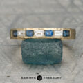 The "Caspian" ring in 14K yellow gold, showcasing alternating blue sapphires and white rectangular gemstones, is displayed on a textured blue stone with the "Earth's Treasury" logo below.