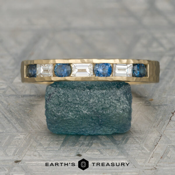 The "Caspian" ring in 14K yellow gold The "Caspian" ring in 14K yellow gold, showcasing alternating blue sapphires and white rectangular gemstones, is displayed on a textured blue stone with the "Earth's Treasury" logo below.