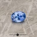 An oval-cut 4.09-carat Cornflower Blue Ceylon Sapphire rests on a textured grey surface. The gemstone's facets catch the light, showcasing its brilliant blue color. The bottom of the image features the text "EARTH'S TREASURY" along with a geometric logo.
