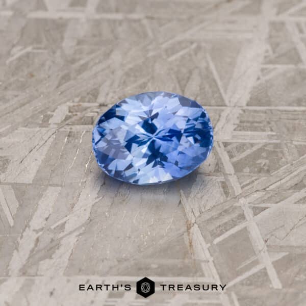 An oval-cut 4.09-carat Cornflower Blue Ceylon Sapphire rests on a textured grey surface. The gemstone's facets catch the light, showcasing its brilliant blue color. The bottom of the image features the text "EARTH'S TREASURY" along with a geometric logo.
