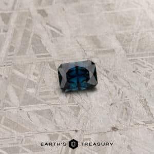 A khaki green gemstone weighing 2.91 carats, cut in a rectangular shape with multiple facets, rests on a gray, textured background. The base of the image features the text "EARTH'S TREASURY" accompanied by a small hexagonal logo.