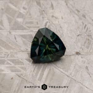 A 2.91-carat Khaki Green Australian Sapphire, with a multi-faceted triangular shape, rests on a textured light-gray surface. The text "EARTH'S TREASURY" is centered below the gemstone.