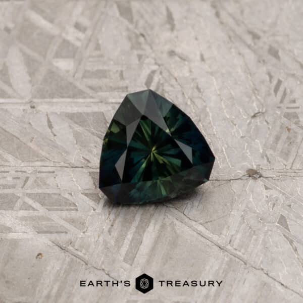 1.72-Carat Dark Green Australian Sapphire (Heated) A 2.91-carat Khaki Green Australian Sapphire, with a multi-faceted triangular shape, rests on a textured light-gray surface. The text "EARTH'S TREASURY" is centered below the gemstone.