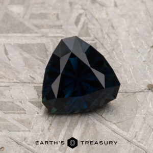 A 1.42-Carat Midnight Blue Australian Sapphire (Heated) with a faceted triangular shape is showcased on a textured gray surface, its deep color catching the light beautifully, with the Earth's Treasury logo featured at the bottom.