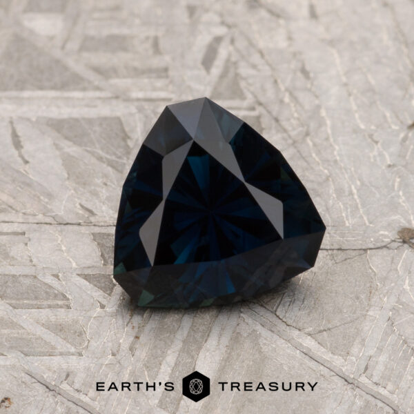 1.42-Carat Midnight Blue Australian Sapphire (Heated) A 1.42-Carat Midnight Blue Australian Sapphire (Heated) with a faceted triangular shape is showcased on a textured gray surface, its deep color catching the light beautifully, with the Earth's Treasury logo featured at the bottom.