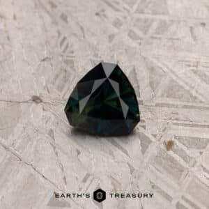 A 2.91-carat khaki green Australian sapphire (heated) featuring a dark, triangular faceted design lies on a textured metallic surface. Below the gem, there is a logo featuring two hexagons and the text "Earth's Treasury.