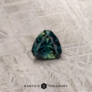 A 2.91-carat khaki green Australian sapphire, expertly cut in a trilliant shape and boasting a rich green-blue hue, rests on a textured surface adorned with geometric designs. The Earth's Treasury logo is prominently displayed at the bottom center of the image.
