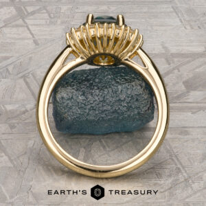 The "Cassandra" ring in 18k yellow gold with a 1.79-carat Montana sapphire is displayed upright on a dark rough stone atop a textured gray surface, with the "Earth's Treasury" logo and text at the bottom.